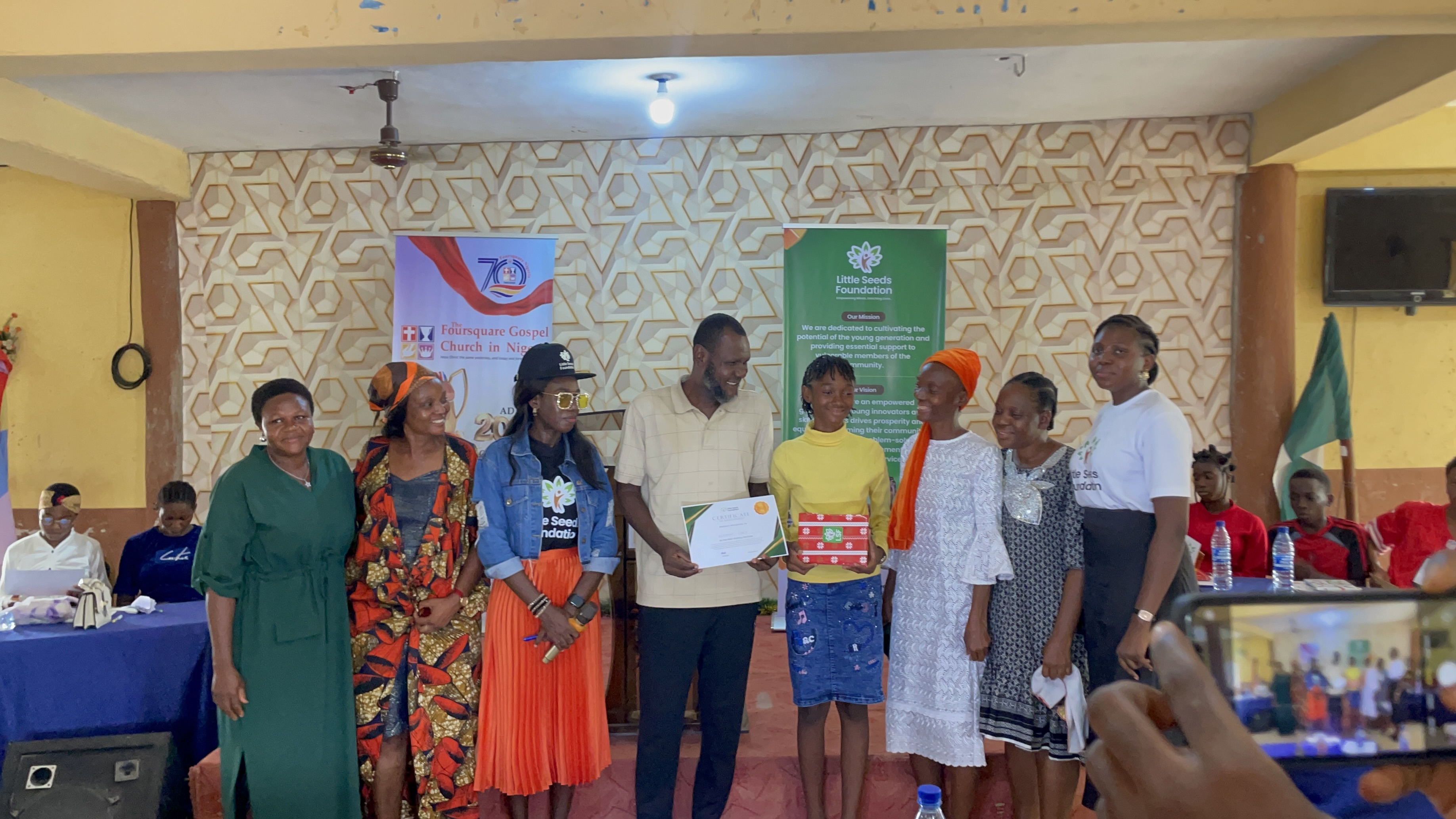 A certificate and prize presented on stage, surrounded by family and supporters